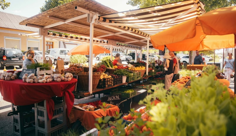 Eine lebhafte Marktatmosphäre mit Menschen, die zwischen bunten Ständen frisches Obst und Gemüse auswählen.